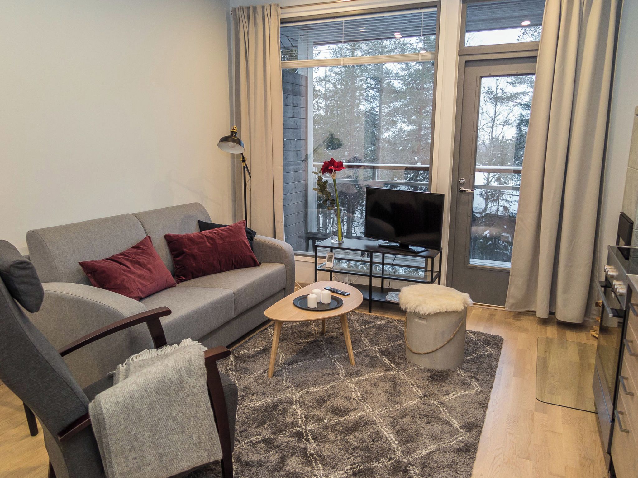 A modern living room with a grey sofa, red cushions, a coffee table with candles, a TV, and a view of trees through large windows.