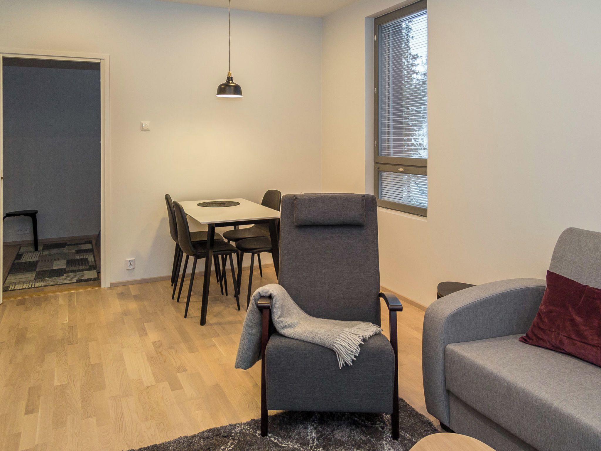 A modern living room with a grey armchair, a grey sofa, a dining table with chairs, and a window with blinds.