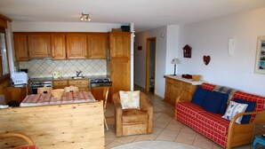 Una cocina con armarios de madera, una mesa de comedor con mantel a cuadros, un sofá con funda de estampado rojo y azul, y una silla roja.