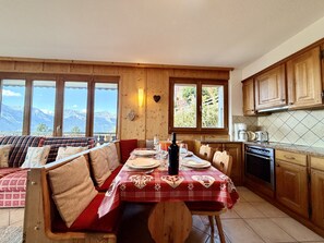 A cozy dining area with a table set for two, wooden cabinets, and a view of mountains through large windows. - Balcons du Soleil Y3 310 - QUIET apartment 6 pers (Veysonnaz)
