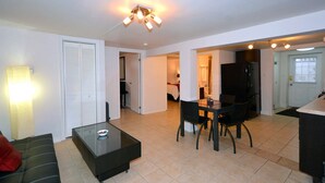A modern living room with a glass coffee table, a dining area with a table and chairs, a kitchen with a refrigerator, and a hallway leading to a bedroom.