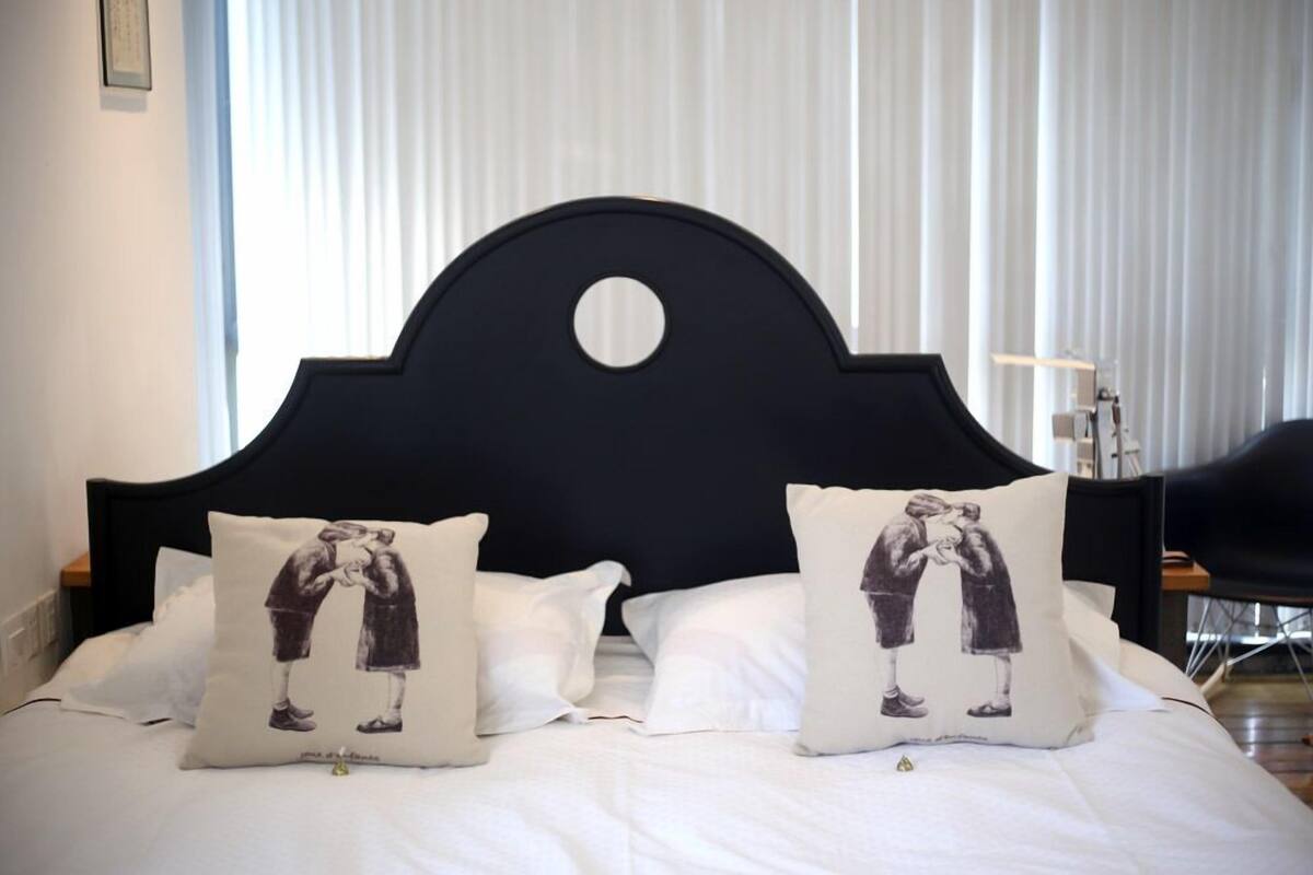 Una cama con cabecera negra, dos almohadas con estampado blanco y negro y ropa de cama blanca.