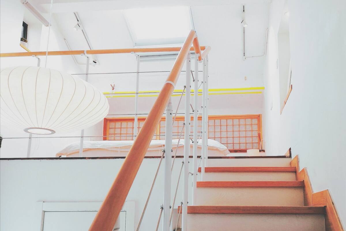 Una escalera con escalones de madera y barandillas de metal, que conduce a una habitación con techo blanco y una lámpara colgante.