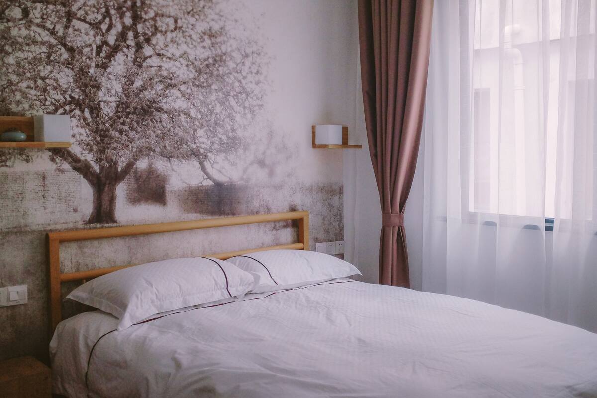 Una cama bien tendida con sábanas blancas, cabecera de madera, estante en la pared con un objeto pequeño y una ventana con cortinas de gasa.