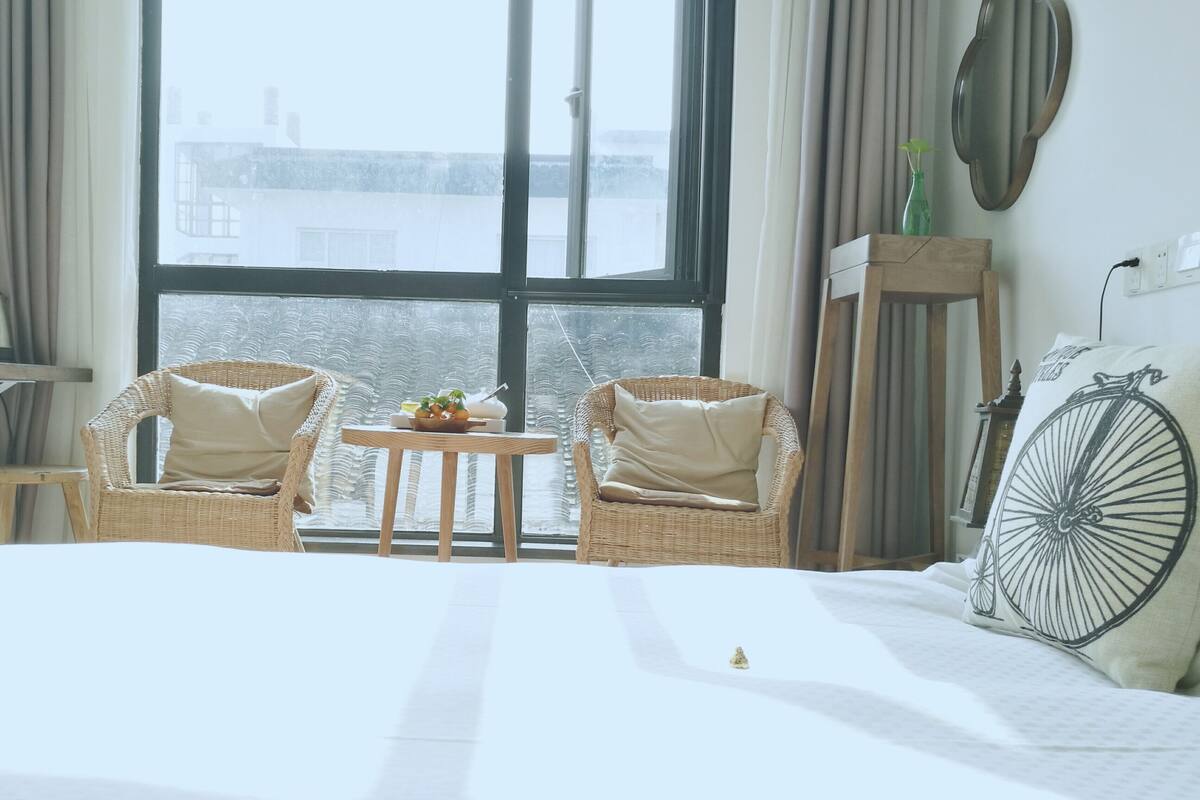 Una habitación de hotel con una cama grande, dos sillas de mimbre, una mesita con frutas, un cojín con forma de rueda de bicicleta y vistas a edificios a través de ventanas amplias.