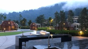 A modern outdoor area with hexagonal structures, a central water feature, and a view of a forested mountain range.