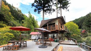 A wooden deck with picnic tables and umbrellas, a wooden building with a balcony, and a forested hillside in the background. - WOOD DESIGN PARK - Campground (Okazaki)