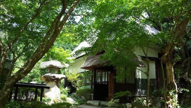 A traditional Japanese-style house with a thatched roof, surrounded by lush greenery and stone steps leading to the entrance. - WOOD DESIGN PARK - Campground (Okazaki)