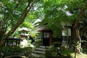 Une maison traditionnelle de style japonais, avec un toit de chaume, entourée d’une végétation luxuriante et des marches de pierre menant à l’entrée.