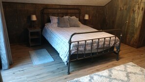 A bedroom with a metal bed frame, a wooden floor, a nightstand with a lamp, and a door to the right. - Ausable River  Cottage (Frederic)