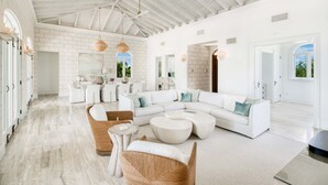 A spacious living area with a white sectional sofa, a round coffee table, and wicker chairs. There is a kitchen area in the background with white cabinets and a dining table. - Exclusive Oceanfront Villa On Taylor Bay Beach! (Providenciales)