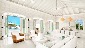 A spacious living room with a white brick wall, a large white sofa, a round coffee table, and a view of a pool and palm trees through open doors. - Exclusive Oceanfront Villa On Taylor Bay Beach! (Providenciales)