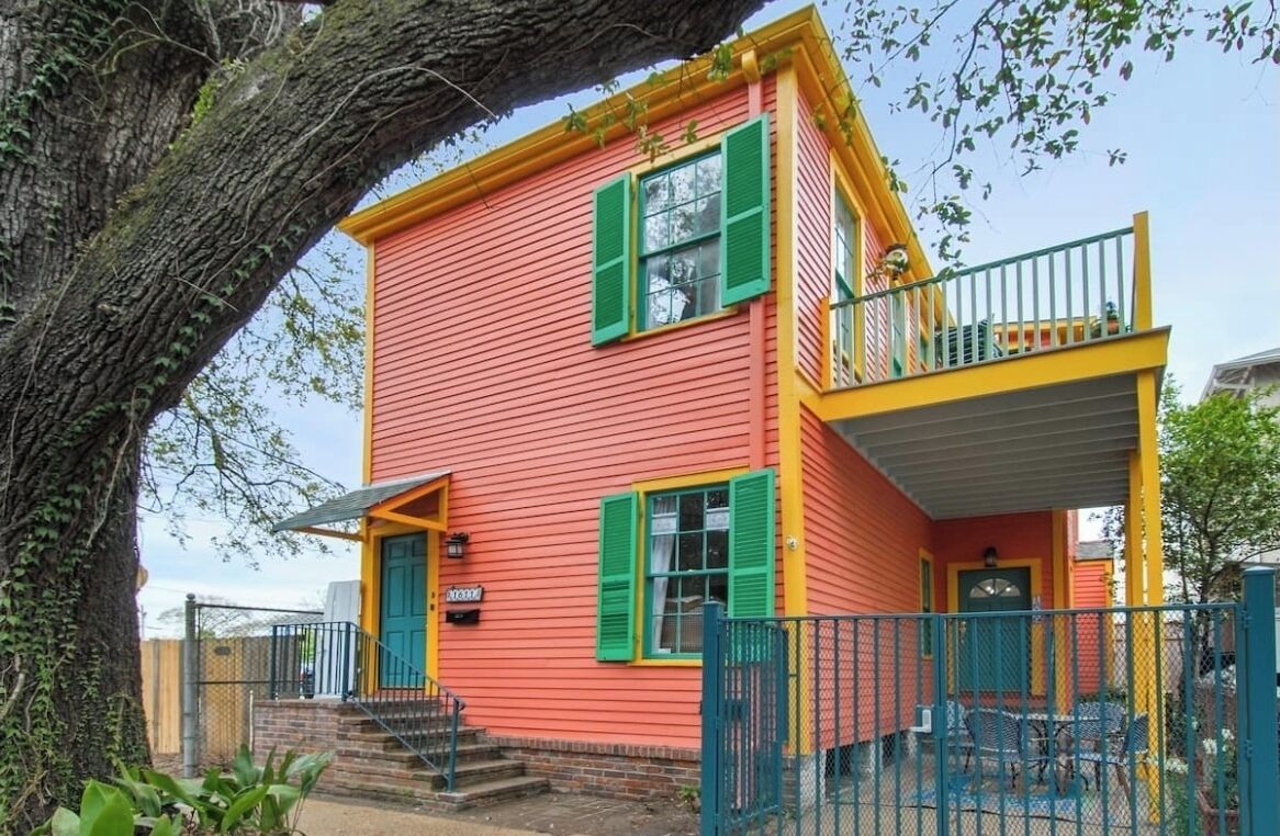 Een twee verdiepingen tellend huis met een rode gevel en groene luiken, een balkon en een omheinde achtertuin.