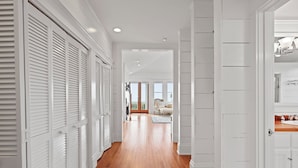 A hallway with white paneling and wooden flooring, leading to a room with a large window and a view of the outdoors. - Beach Lawn Cottage: Premier Oceanfront Escape | Amazing Views | Steps to Beach | Beachfront Pools (Saint Simons Island)