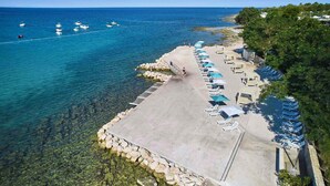 Vue aérienne d’une plage avec une eau d’un bleu limpide, une promenade en béton et des rangées de chaises longues.