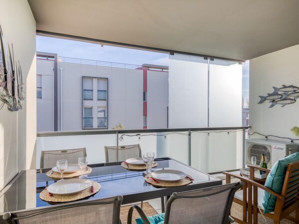 A dining area with a glass table set for four, chairs, and a view of buildings through large windows. - Le Newport by Interhome (Fréjus)