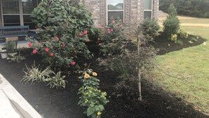 Una casa con fachada de ladrillo, un porche cubierto y un jardín con diversas plantas y flores.