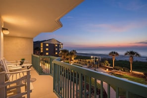 A balcony with a view of the ocean at sunset, outdoor seating, and a building in the background.