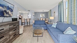 A modern living room with a blue sofa, a glass coffee table, and a flat-screen TV on a wooden cabinet. - Avista Resort 720 - 3 Bedroom Oceanfront Condo with Lots of Amenities! (NORTH MYRTLE BEACH)