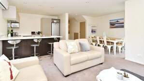 A modern living room with a sofa, a dining area with a table and chairs, a kitchen with a refrigerator and bar stools, and a painting on the wall. - Hagley Apartment (Christchurch)