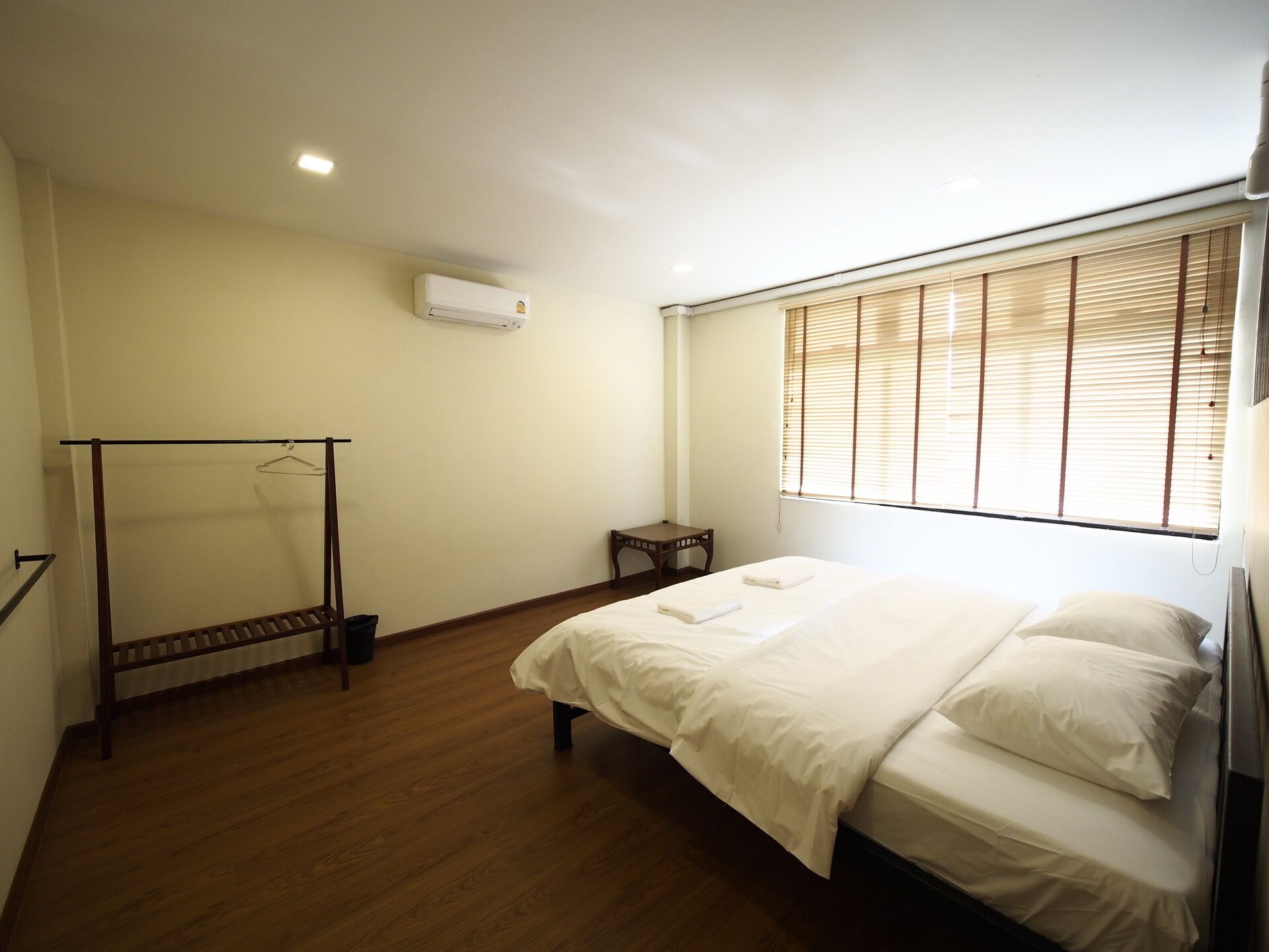 A bedroom with a bed, a wooden floor, a bedside table, a hanging rack, and a window with blinds.