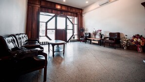 A spacious lobby with a leather sofa, a wooden coffee table, and a seating area with chairs. A large window with a geometric design allows natural light to enter. - More than Sleep Hostel Pak Chong - Khao Yai (Pak Chong)