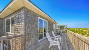 Une terrasse en bois avec des chaises blanches, une porte vitrée et une vue sur la plage.