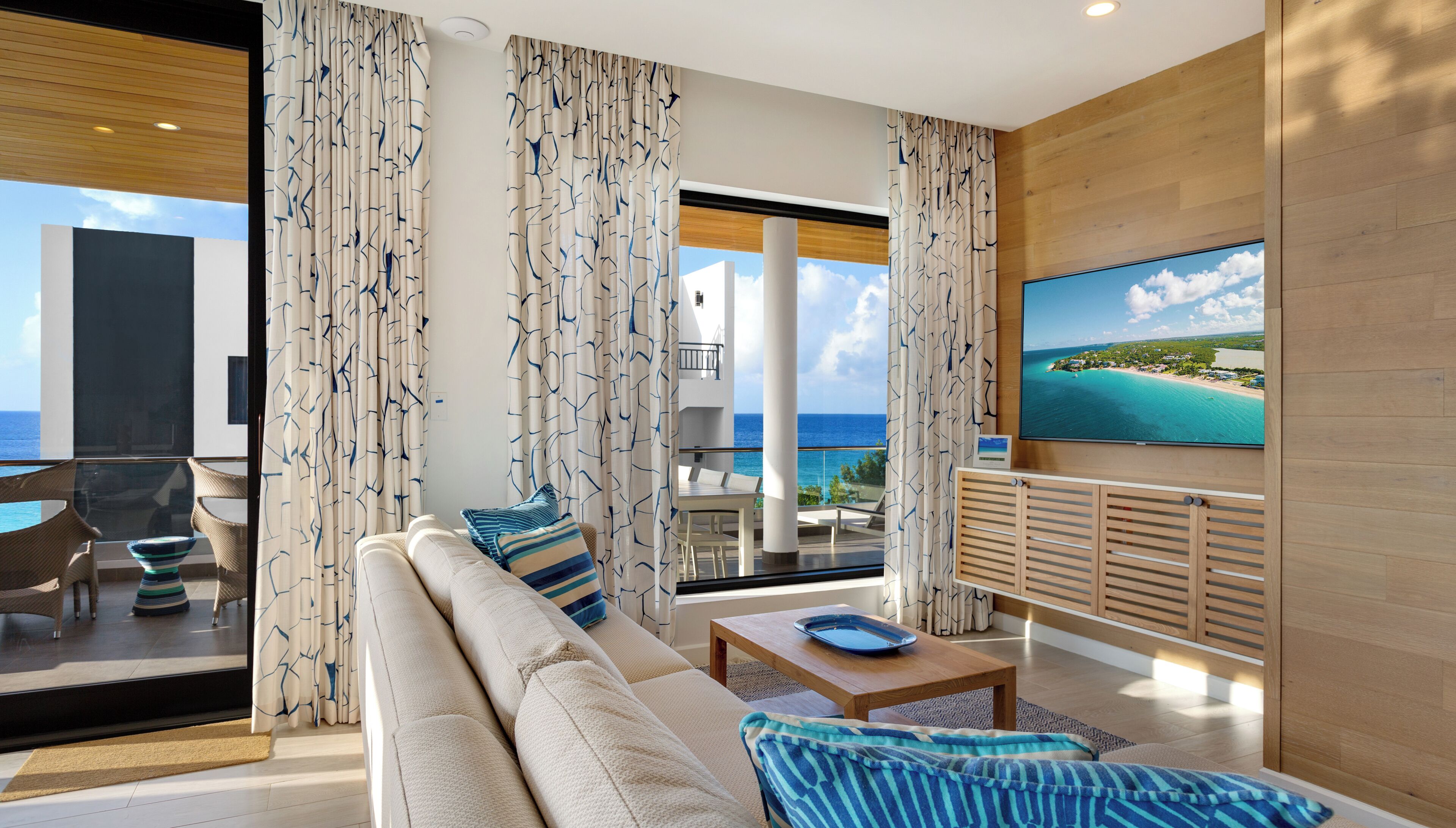 A modern living room with a sofa, coffee table, and TV, overlooking a balcony with a scenic ocean view.