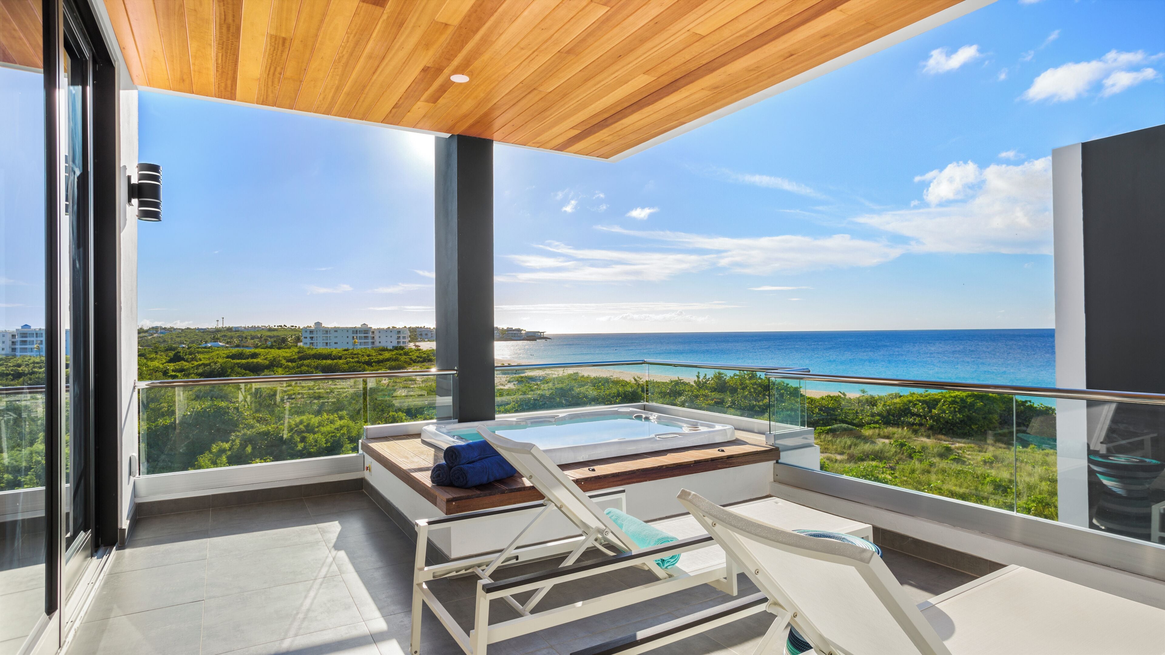 A modern balcony with a glass railing, a hot tub, and lounge chairs overlooking a beach and ocean.