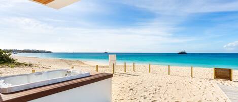 A beach view from a balcony with a hot tub, wooden deck, and a sandy beach.