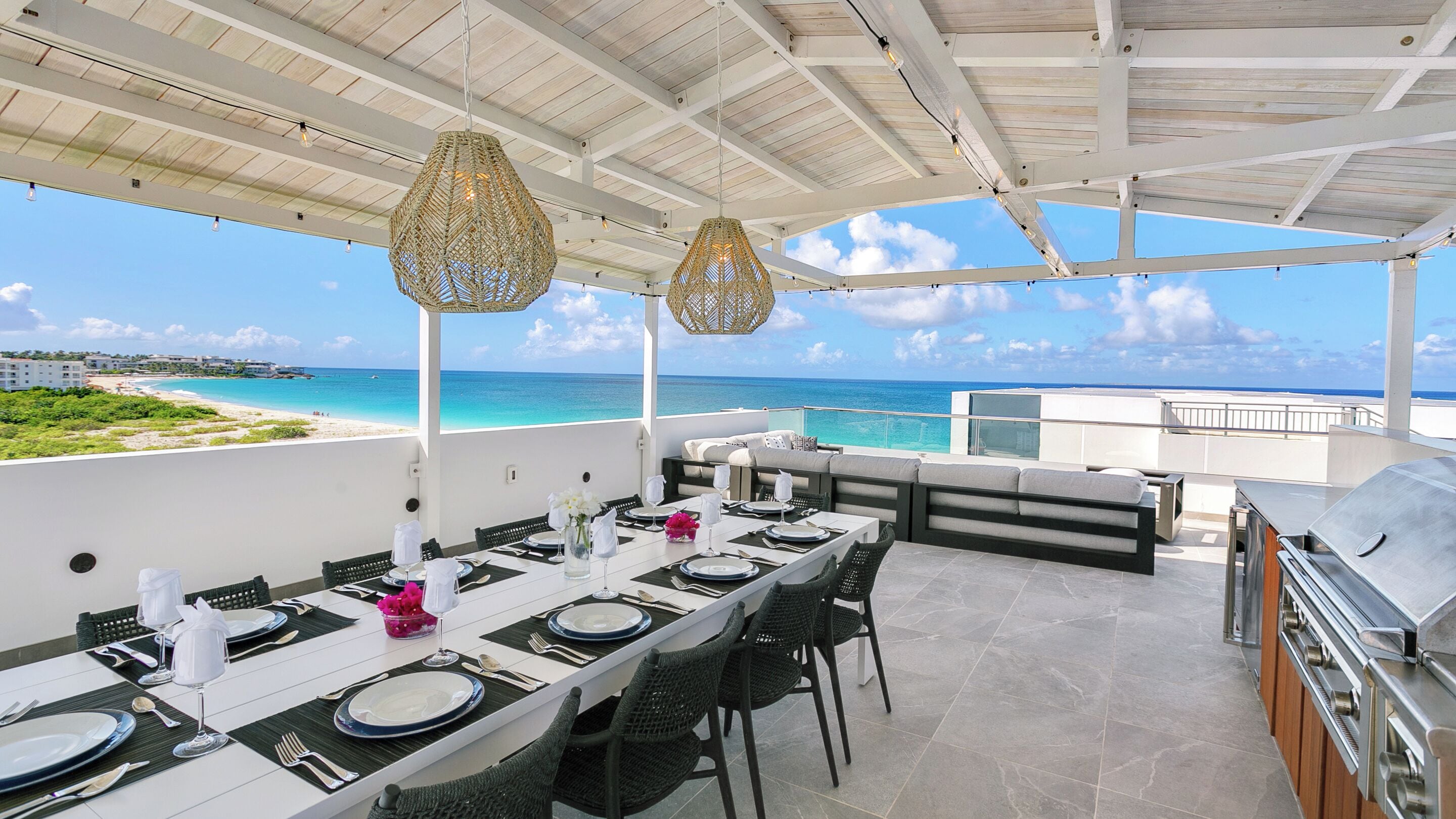 An outdoor dining area with a view of the beach and ocean, a grill, and a covered seating area.