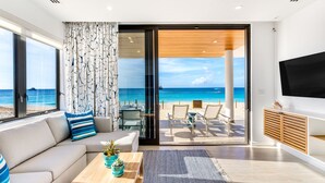 A modern living room with a large window overlooking a beach, a sofa, a coffee table, and a TV mounted on the wall. - Tranquility Beach Anguilla (West End Village)