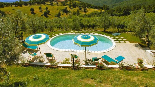 Ein Poolbereich mit zwei runden Pools, umgeben von Liegestühlen und Sonnenschirmen, vor der Kulisse grüner Hügel und einem klaren Himmel.
