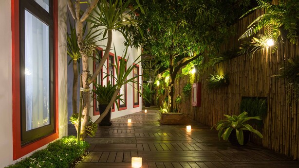 A wooden pathway flanked by lush greenery and potted plants, leading to a building with a red-framed window. - De Sarann Villa (Siem Reap)