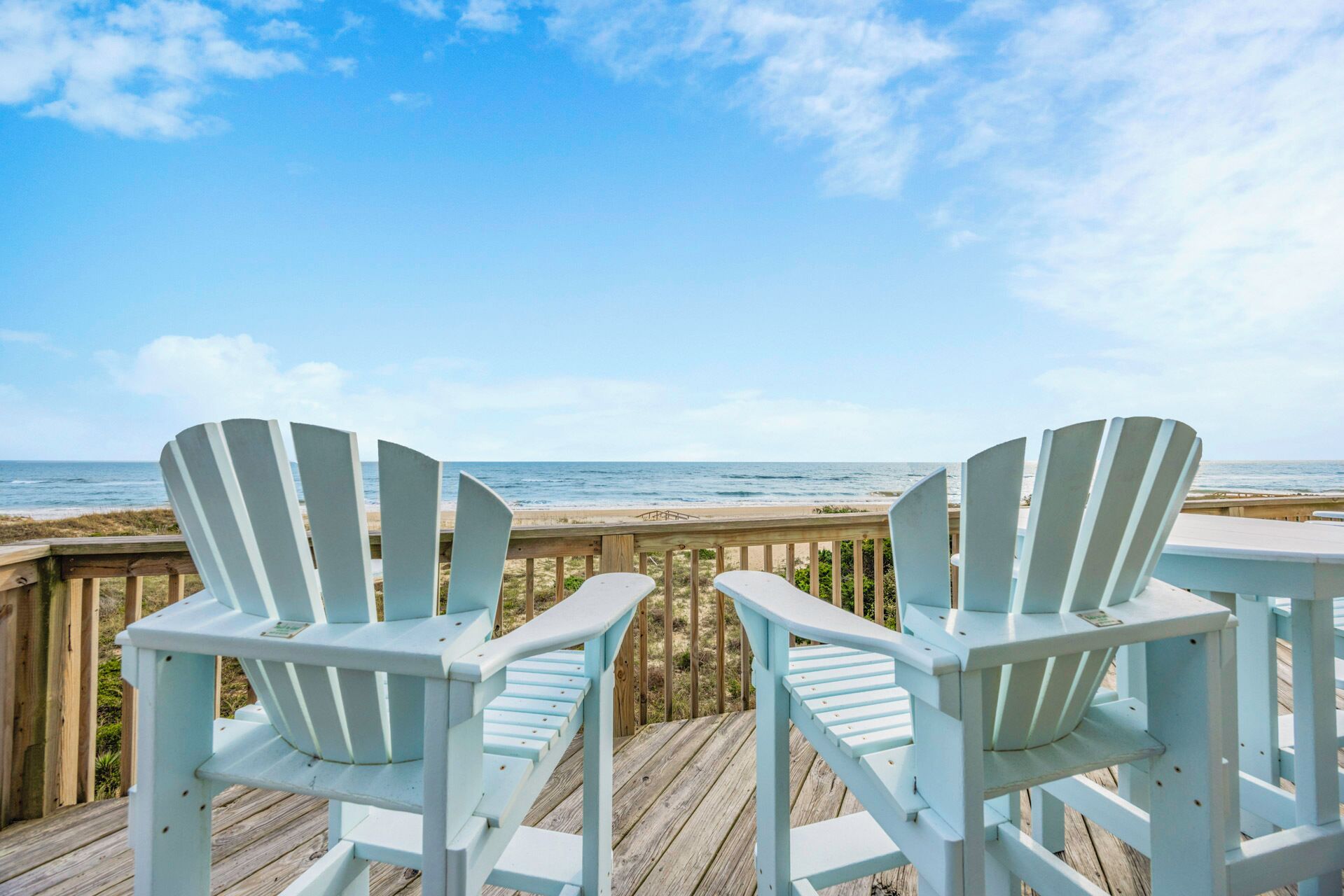 Two white Adirondack chairs on a wooden deck overlooking a beach with the ocean in the background.
