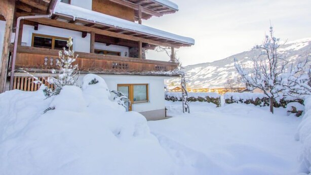 Un chalet en bois à deux étages, doté d’un balcon, entouré de terrain enneigé et avec des montagnes en arrière-plan.