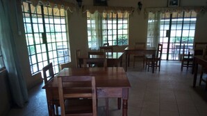 A dining area with wooden tables and chairs, large windows with curtains, and a tiled floor. - Philadelphia Retreat and Conference (Mururi)