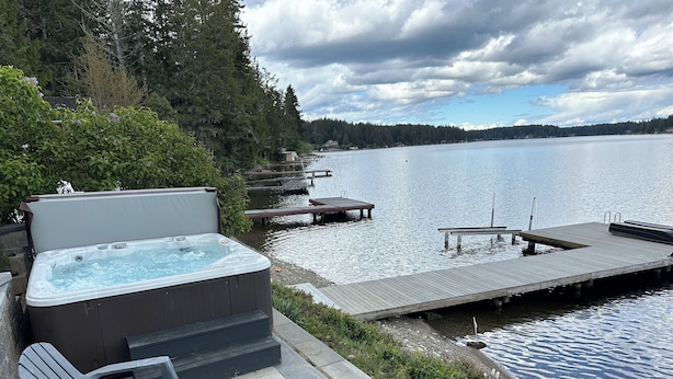A hot tub, outdoor chairs, and a wooden dock extending into a lake with a forested shoreline.
