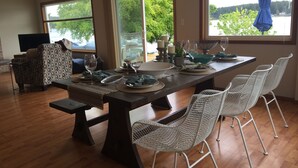 A dining area with a long table set for six, surrounded by wicker chairs, with a view of trees and a body of water through large windows.