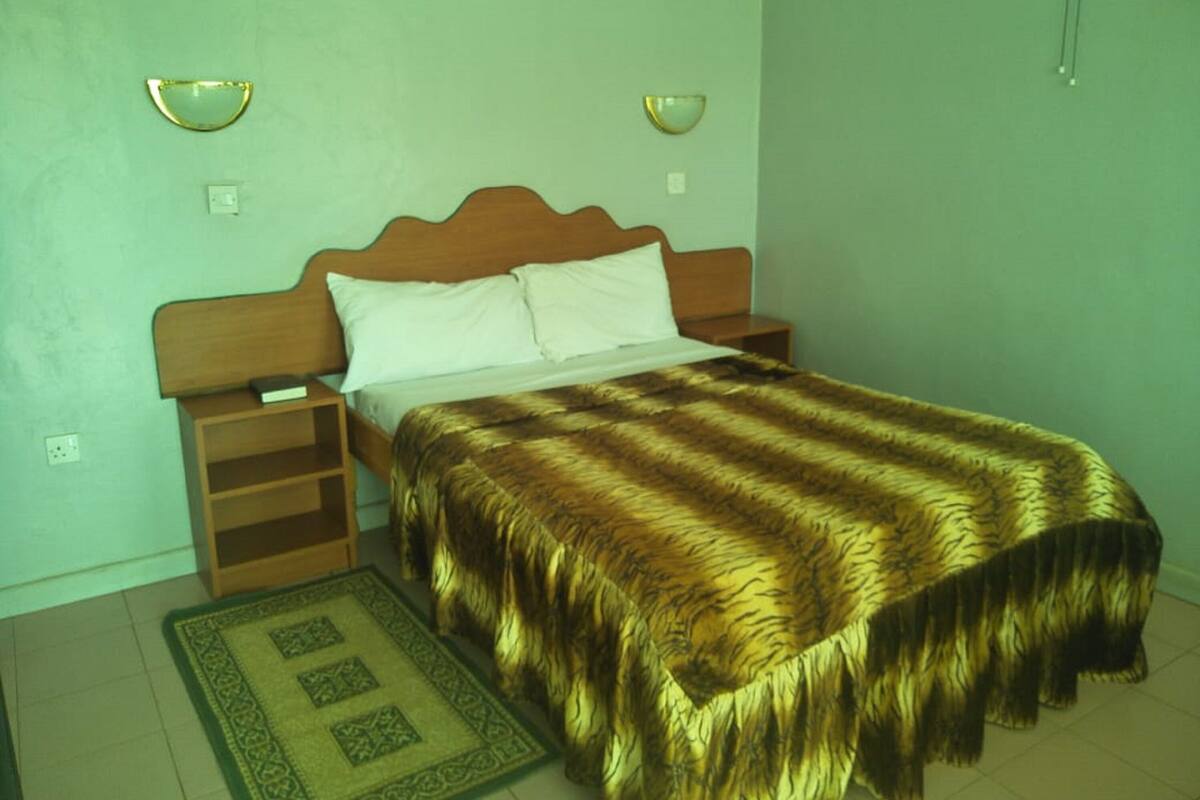 A bedroom with a wooden bed, white pillows, a brown shaggy bedspread, a bedside table, a nightstand, and a green patterned rug.
