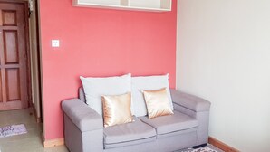 Een grijze bank met twee kussens tegen een roze muur, een wit schapf boven en een houten deur links.