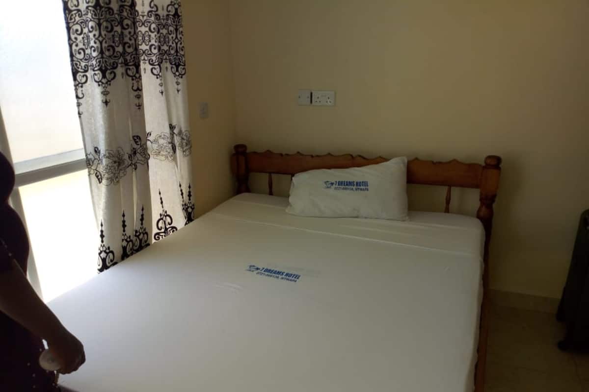 A hotel room with a bed, a pillow with the hotel name, a window with patterned curtains, and a wall socket.