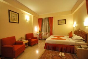 A hotel room with a bed, two armchairs, a small table with a vase, and a nightstand with a phone. - The Senator Hotel (Kolkata)