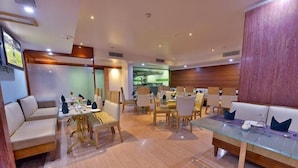 A modern hotel dining area with a glass-top table, cushioned seating, and a view of the kitchen. - The Senator Hotel (Kolkata)