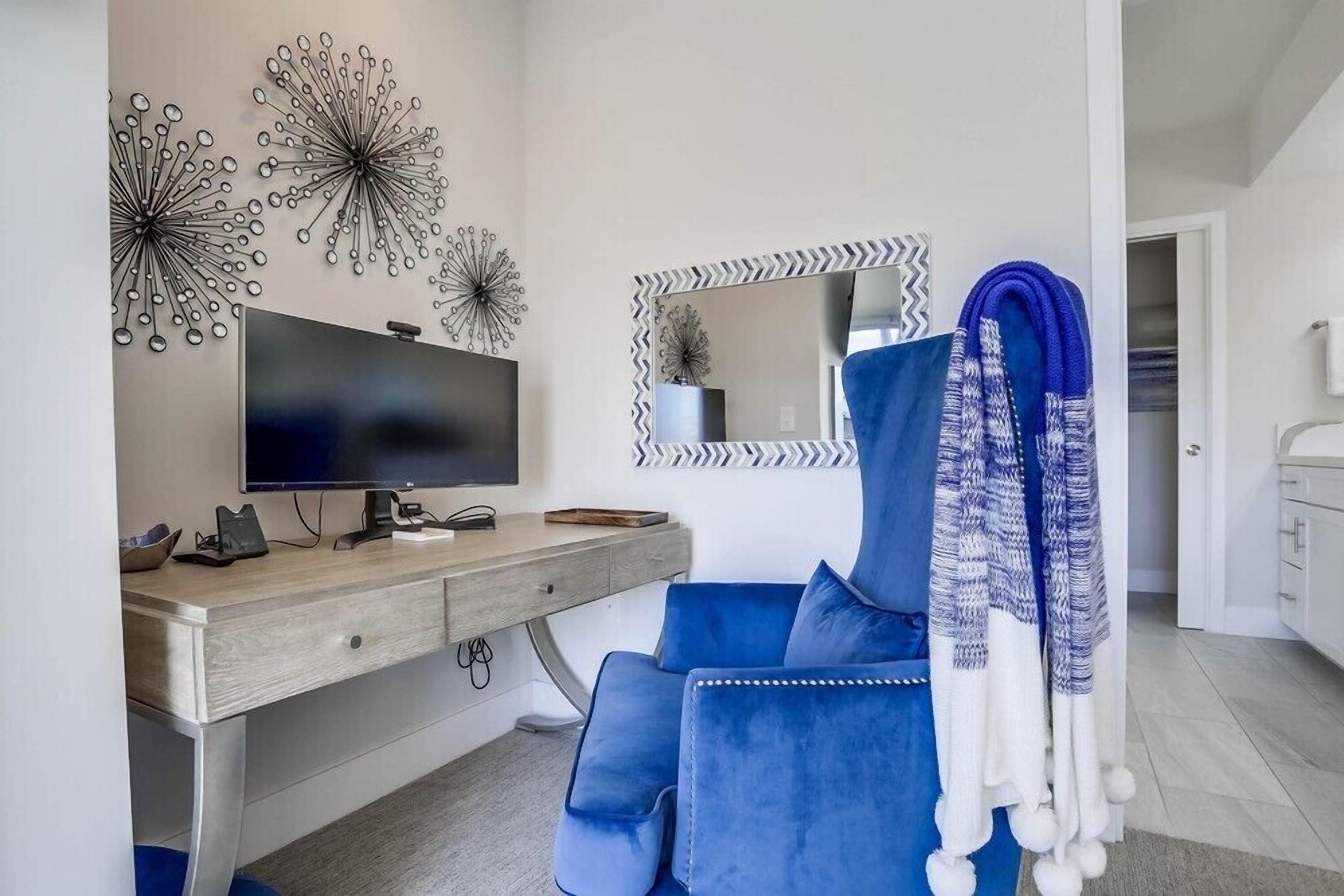 Ein modernes Schlafzimmer mit einem Holztisch, einem großen Flachbildfernseher, einem blau gepolsterten Sessel, Wanddekoration und einem Spiegel mit Zickzackrahmen.