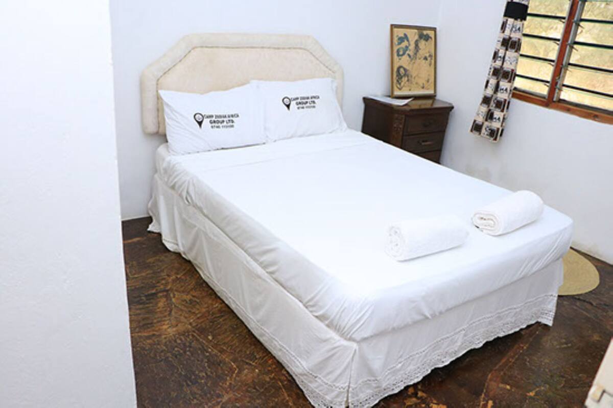 Una cama bien tendida con sábanas y almohadas blancas, una mesita de noche de madera con una lámpara, y una ventana con vistas al exterior.