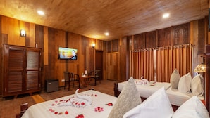 Una habitación de hotel con paneles de madera, dos camas con sábanas blancas y pétalos de rosa, un televisor en la pared y una mesa pequeña con sillas.