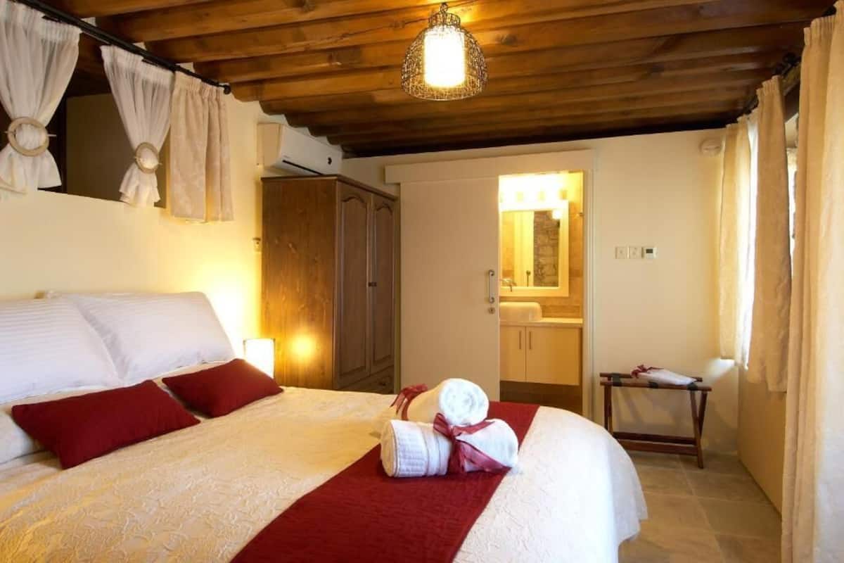 A bedroom with a wooden ceiling, a bed with white linens and red pillows, a bedside table with a towel, and a mirror above the sink.
