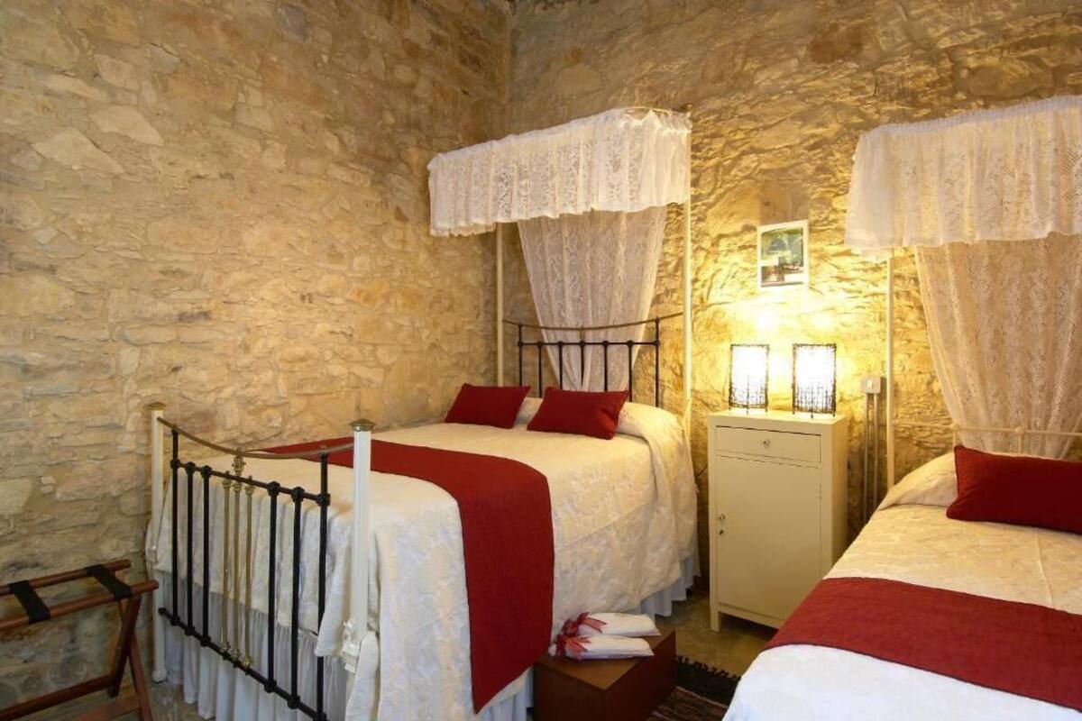 A room with stone walls, two beds with white and red bedding, and a nightstand with a lamp.