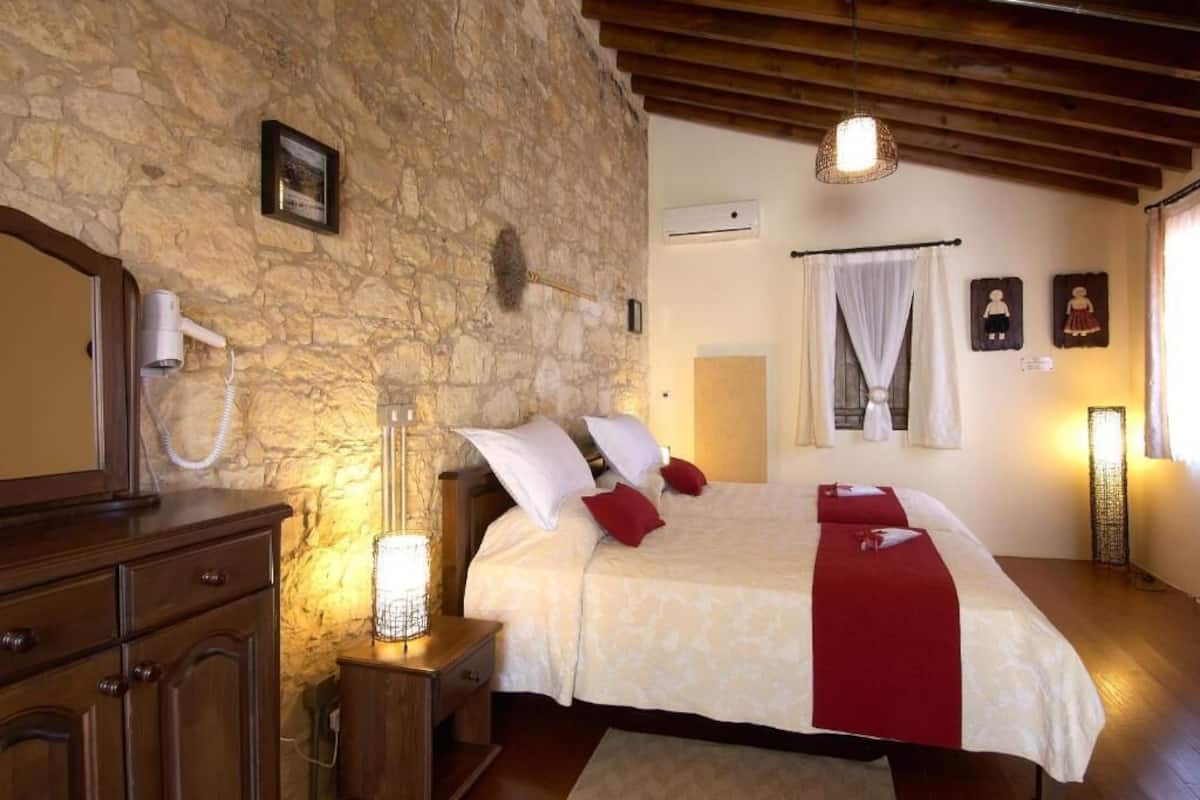 A bedroom with a stone wall, wooden ceiling beams, a bed with white and red bedding, a bedside table with a lamp, and a dresser.